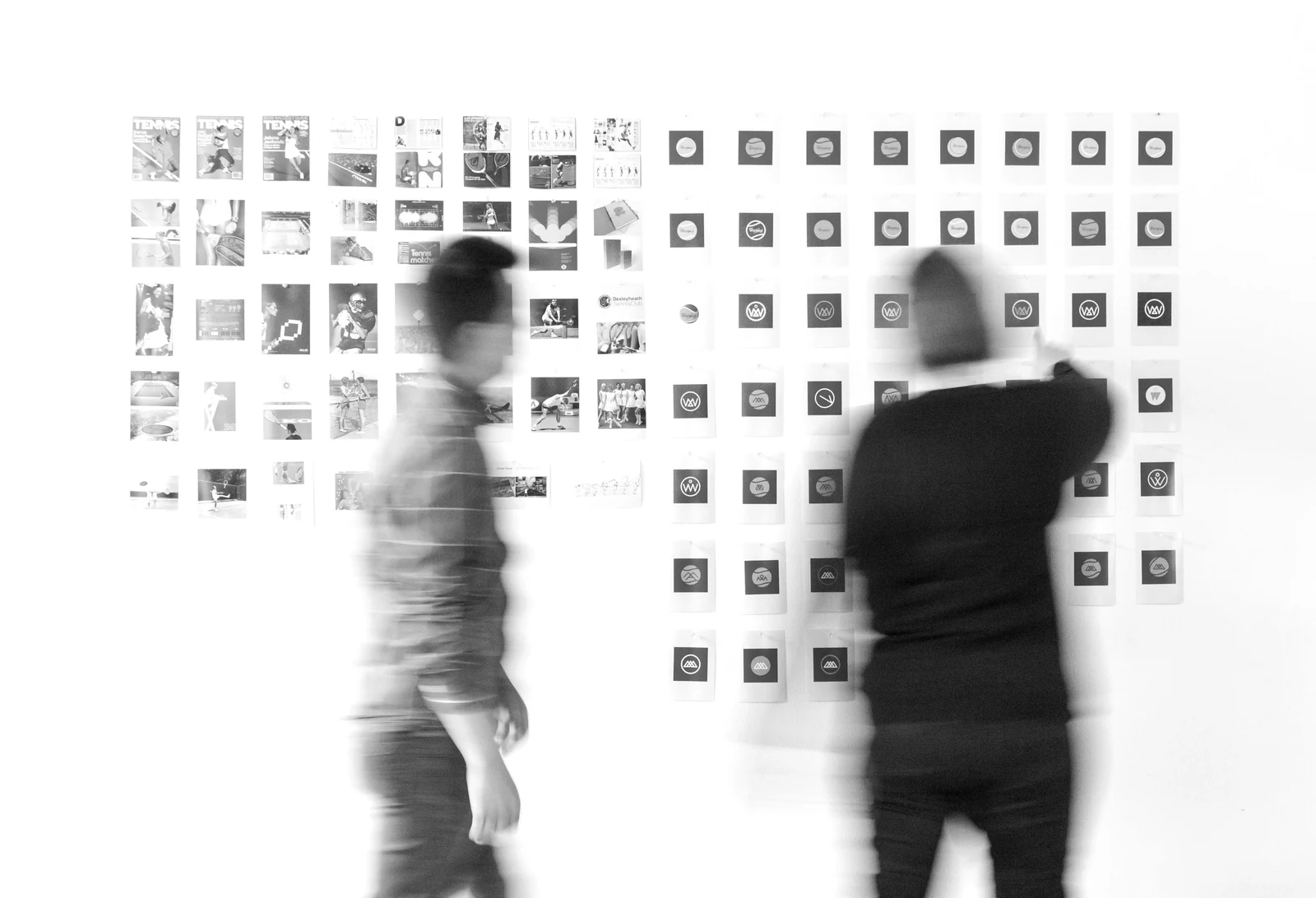 Two blurred people standing before a wall covered in small printed images and logos. Photograph accompanying Velaharra’s artist portfolio for representation guide.